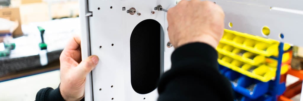 Hands assemble a white sheet metal panel with holes, next to a rack of small bins