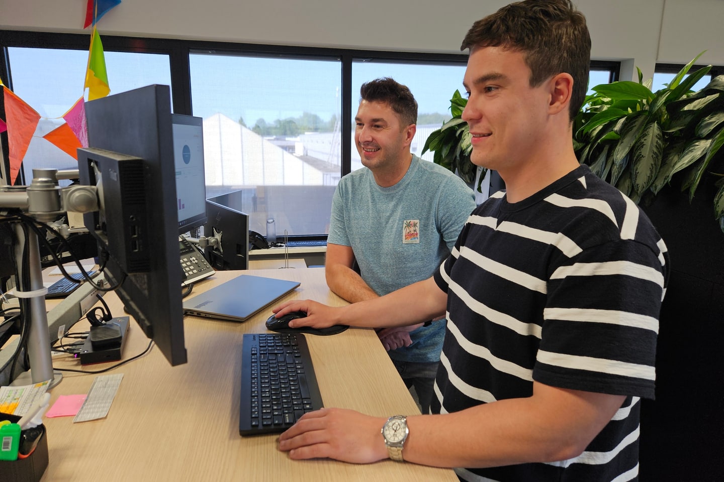 Twee collega's van de Orderdesk aan een hoog bureau met beeldschermen, met uitzicht op de fabriek