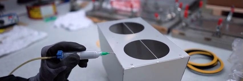 Hand mit Handschuh trägt mit einer Spritze Kleber auf ein Aluminiumgehäuse mit zwei runden Öffnungen auf
