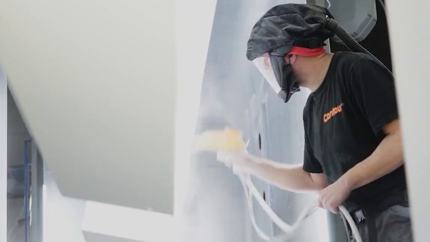 Medewerker met gelaatsscherm spuit poeder op een wit plaatwerkdeel in de spuitcabine