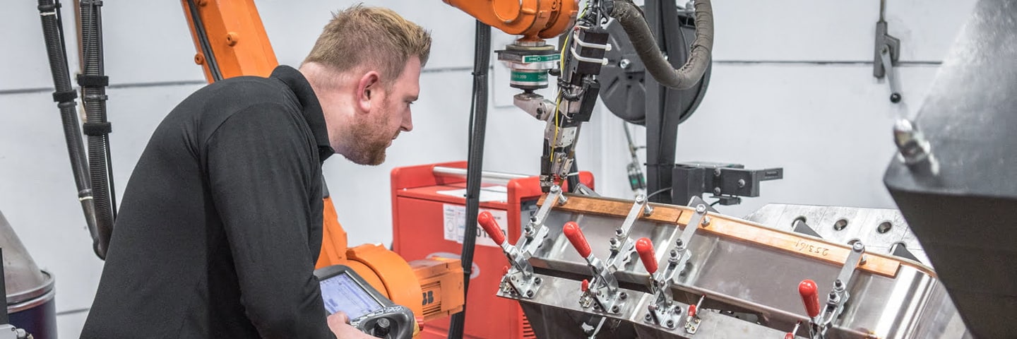 Operator programming an ABB welding robot with the teach pendant; the sheet metal part is clamped in a welding jig