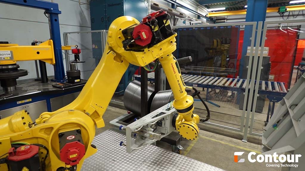 Gele robotarm met grijper bij een machine, met een rollenbaan en een lasscherm op de achtergrond