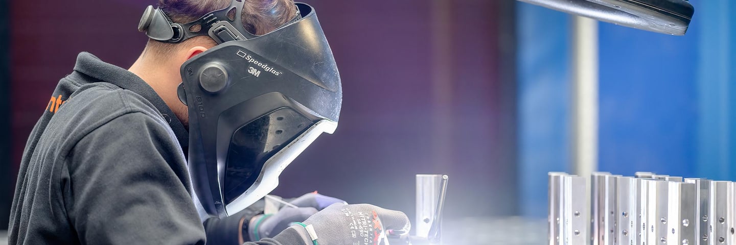 Welder with helmet TIG welding stainless steel parts, a row of welded stainless parts on the right