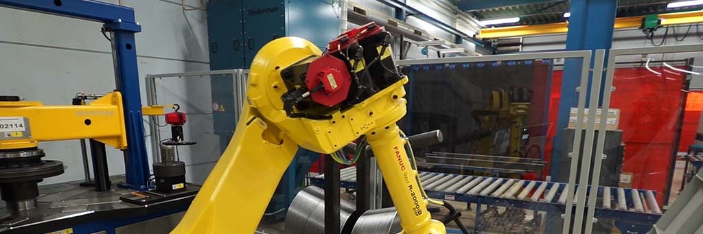 Yellow robot arm with gripper at a machine, with a roller conveyor and a welding screen in the background