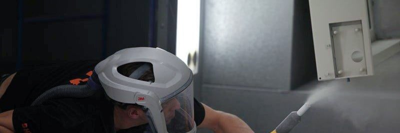 Powder coater in a respirator helmet sprays powder onto a sheet metal part in the booth