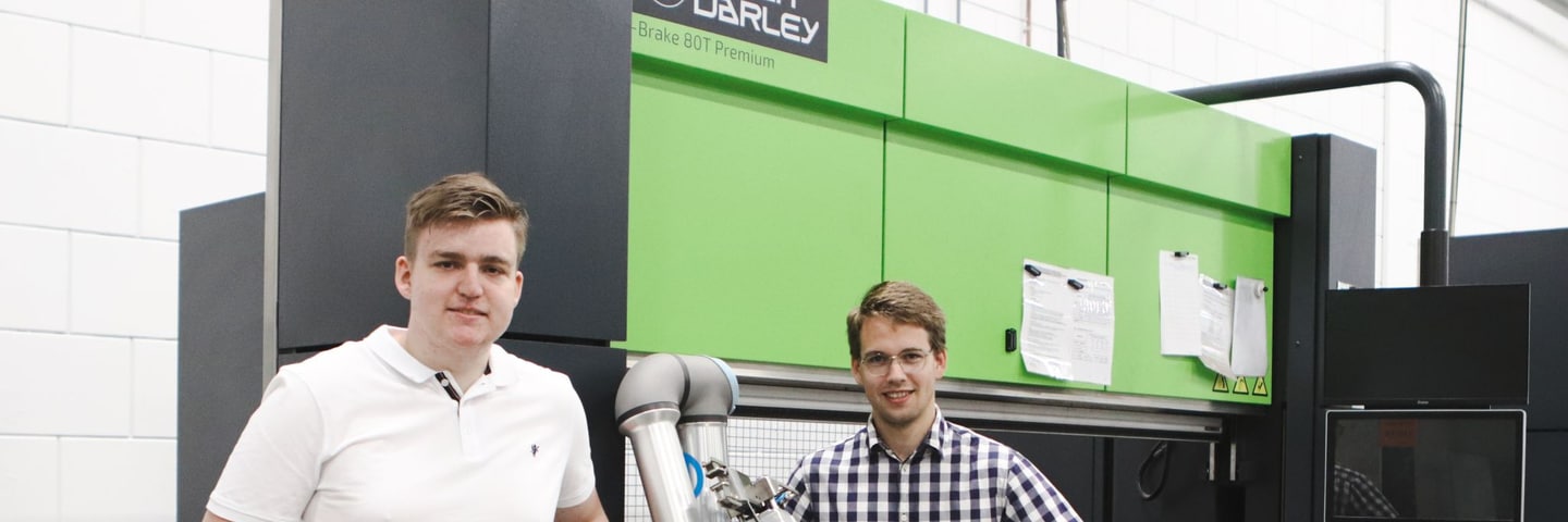 Zwei neue Studenten stehen neben einer Abkantpresse mit Cobot