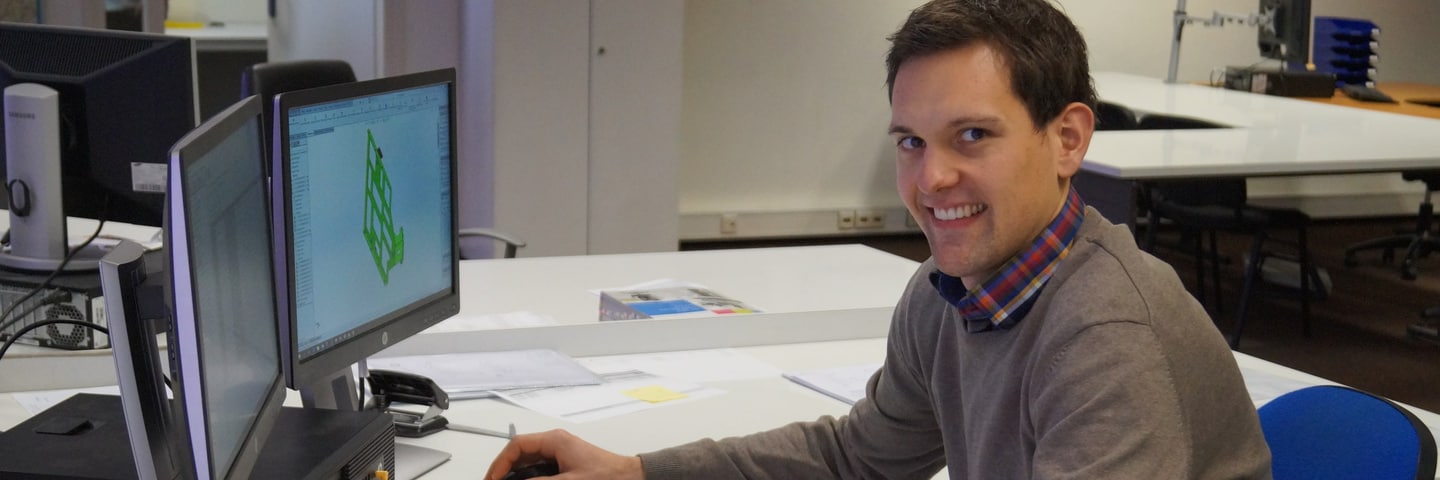 Collega zit aan een bureau met twee beeldschermen en kijkt lachend in de camera
