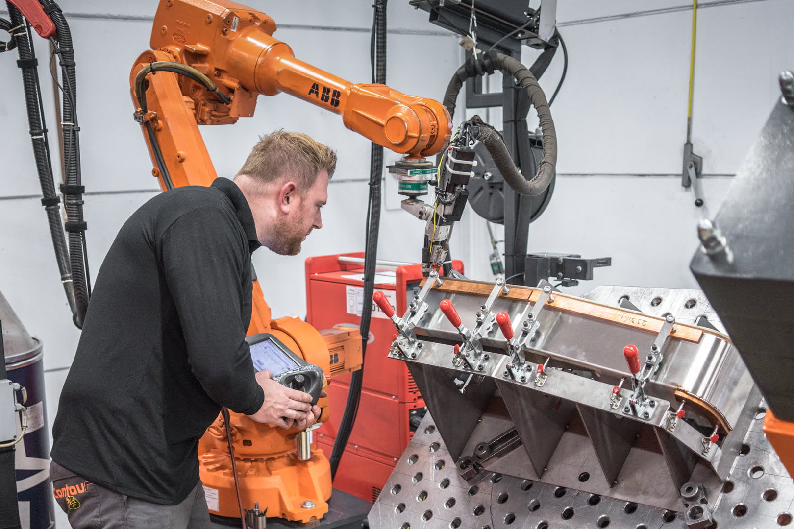 Operator programming an ABB welding robot with the teach pendant; the sheet metal part is clamped in a welding jig