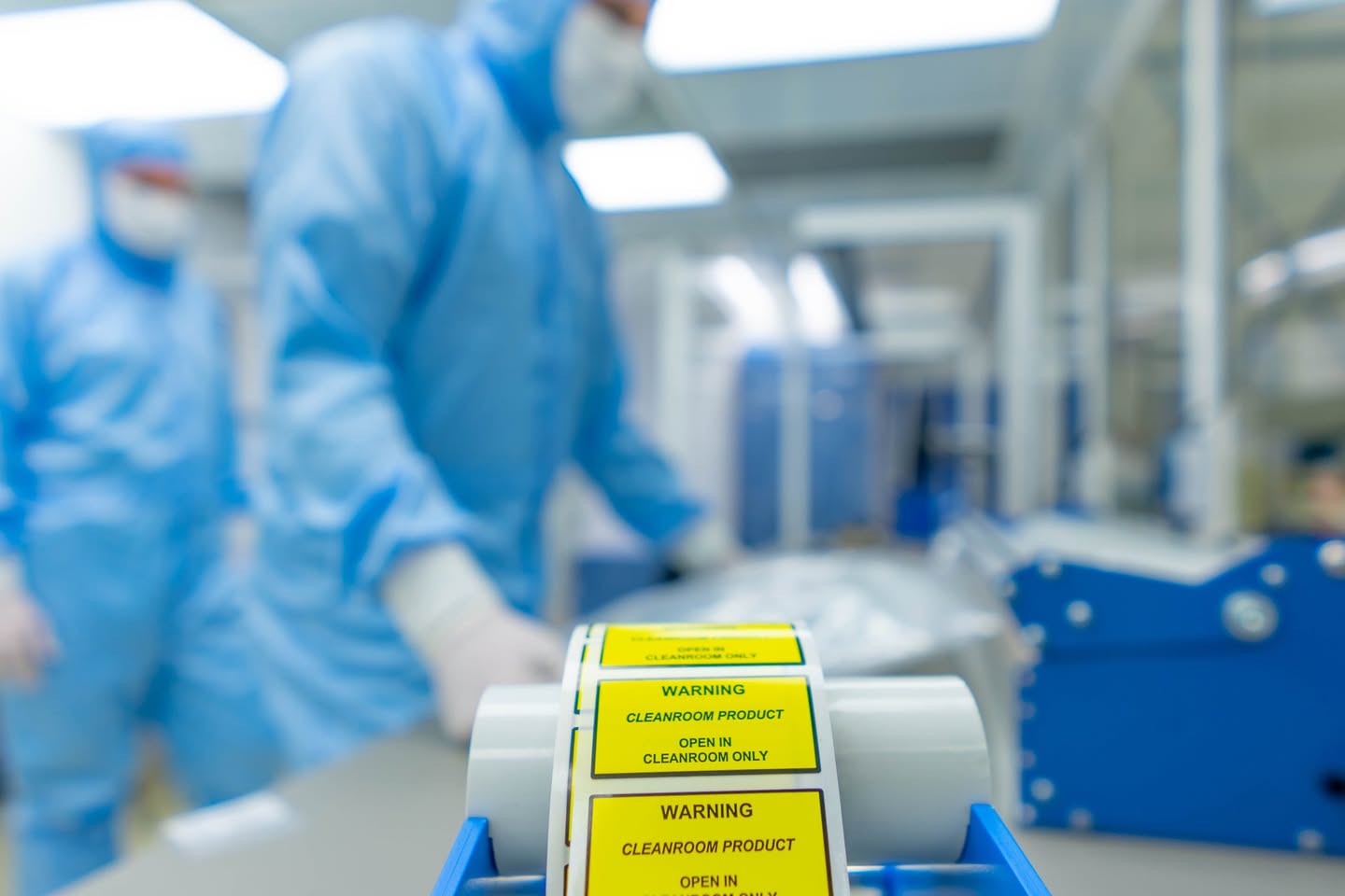Rol gele cleanroom-etiketten op de voorgrond, met twee medewerkers in blauwe cleanroomkleding