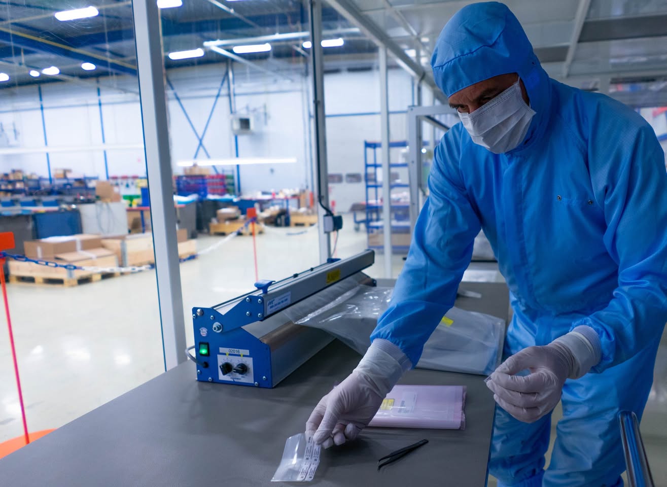 Medewerker in cleanroomkleding verpakt een onderdeel in een zak naast een foliesealer