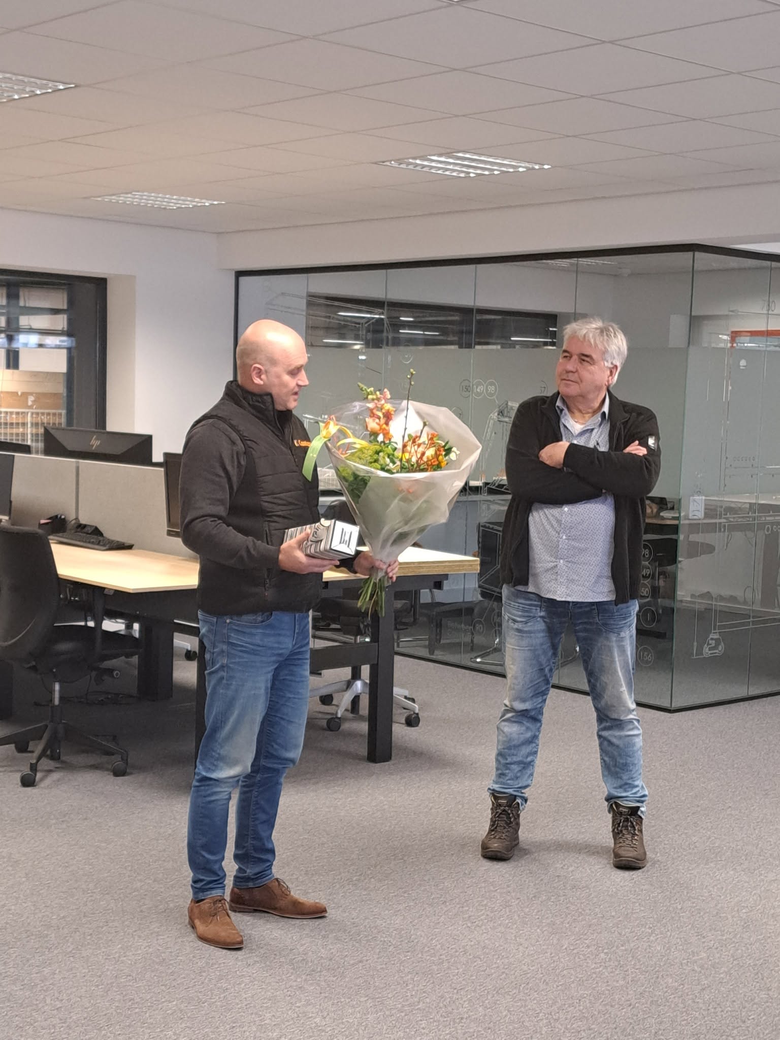 Een collega spreekt Wim toe met een bos bloemen in het kantoor