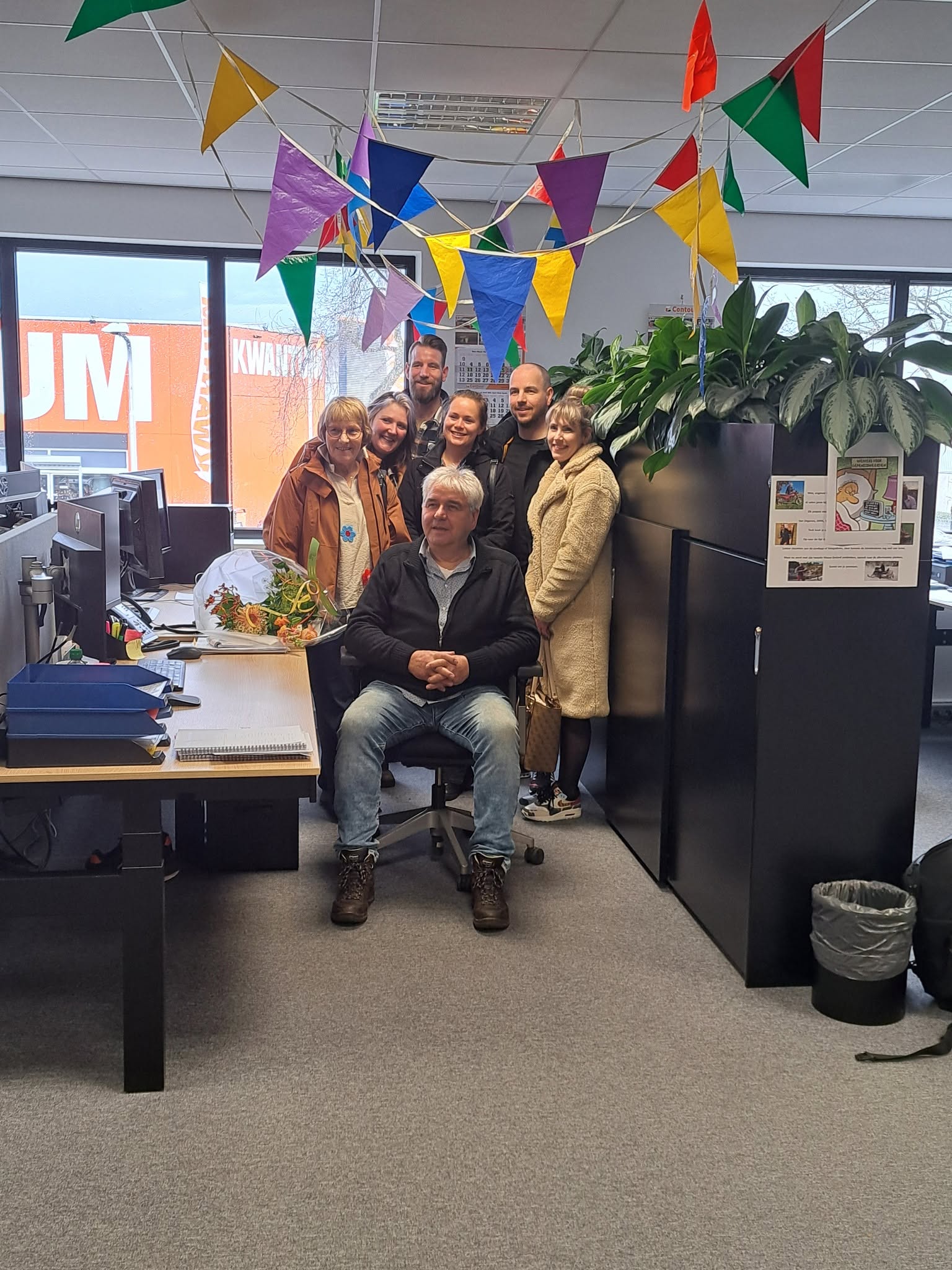 Wim op een bureaustoel met collega's om hem heen en vlaggetjes aan het plafond